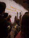 A group of people in front of a wall watching a projection. The projection shows two shadowy silhouettes in the process of writing the phrase “Welcome to the Canadian Museum for Human Rights” in English and French.