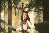 Une artiste de drag porte un costume de cosplay avec de longs cheveux bruns, de grands bois de cerf, ainsi qu’un foulard, une jupe et des gants en forme de feuilles vertes.