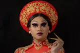 Une artiste de drag porte des vêtements culturels vietnamiens avec une coiffe et une robe longue rouge et or assorties.