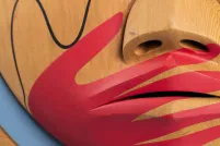 Un plan rapproché d’une boîte en bois, sur laquelle est sculpté un visage dont la bouche est recouverte d’une main peinte en rouge.