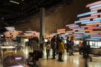 Un grand groupe de personnes dans une galerie dont un mur est recouvert de panneaux colorés et de photos montrant une chronologie. 