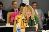 Une femme blonde parlant dans un microphone. Elle est assise et d'autres personnes sont assises derrière elle.