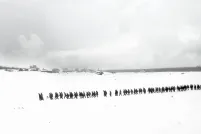 Une photo en noir et blanc d’un paysage plat et enneigé. On voit des arbres et des petits bâtiments au loin. Une longue file de plus de 100 personnes marchent dans la neige.