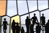 Dix-huit personnes, certaines assises, certaines debout, placées en rangée le long d’un mur de fenêtres aux formes irrégulières.