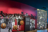 Une galerie muséale avec un grand char d'assaut jaune sortant d'un mur et des couvercles rouillés de poubelle accrochés au mur d'en face.
