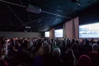 Un auditoire assis regarde trois grands écrans dans une salle dont l'éclairage est tamisé.