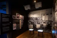 Une exposition muséale montrant une photo en noir et blanc d’enfants assis à des pupitres placés en rangées. Deux pupitres semblables à ceux qu’on voit dans la photo sont placés au centre de l’exposition. Sur un panneau de texte, on peut lire « Enfances oubliées ».
