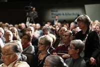 Une salle remplie de personnes assises. Une femme est debout et tient un microphone portable.
