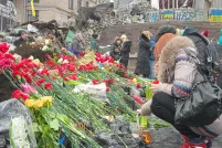 Deux personnes déposant des fleurs rouges à un mémorial. En arrière-plan, une douzaine d’autres personnes se tiennent devant des débris, comme des roches, des feuilles de métal et des barrières de ciment peintes avec des drapeaux ukrainiens bleu et jaune et des lettres.