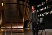 Une jeune femme parle devant un mur noir qui dit, en grosses lettres blanches, « Tous les êtres humains naissent libres et égaux en dignité et en droits ». Il y a un théâtre circulaire à l’arrière-plan qui ressemble à un grand panier tressé.
