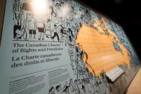 Sur le mur d’une exposition muséale figure une fresque illustrée représentant divers personnages et le titre « The Canadian Charter of Rights and Freedoms / La Charte canadienne des droits et libertés ». La fresque passe d’illustrations noires représentant des personnes à une partie orange où sont énoncés, en anglais et en français, les droits et libertés fondamentaux, qui prennent la forme du Canada.