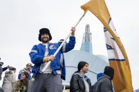 Un homme souriant dans une foule, tenant un grand drapeau jaune avec un « W » bleu et blanc au centre. L’homme porte une veste bleue des Blue Bombers de Winnipeg et se tient devant le Musée canadien pour les droits de la personne.