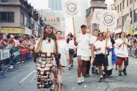 Un groupe de personnes portant des pancartes sur lesquelles on peut lire « 2-Spirited People of the 1st Nations » marchent au centre d’une rue de la ville, tandis que des gens regardent derrière des barrières de part et d’autre de la rue.