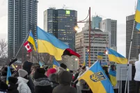 Une foule de personnes emmitouflées se tient debout dans une rue de ville pendant l’hiver, tenant de multiples drapeaux ukrainiens bleus et jaunes ainsi qu’un drapeau canadien. Le rassemblement semble être une manifestation en appui à l’Ukraine. De grands édifices à bureaux et des feux de circulation sont à l’arrière-plan sous un ciel couvert.