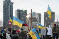 Une foule de personnes emmitouflées se tient debout dans une rue de ville pendant l’hiver, tenant de multiples drapeaux ukrainiens bleus et jaunes ainsi qu’un drapeau canadien. Le rassemblement semble être une manifestation en appui à l’Ukraine. De grands édifices à bureaux et des feux de circulation sont à l’arrière-plan sous un ciel couvert.