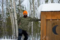 Un homme sikh-canadien, souriant, portant un pull vert, un pantalon bleu et marron, des bottes et un turban orange, adossé à une cabane avec un croissant de lune.