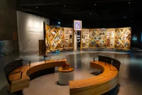 Exposition muséale présentant un grand mur de bois incurvé et autoportant sur lequel sont fixés de nombreux petits objets disposés en losanges et en rectangles. Au premier plan, des bancs circulaires créent un espace accueillant où les gens peuvent regarder et contempler l’exposition.