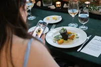 Une femme à la peau claire et aux longs cheveux bruns utilise son téléphone portable pour prendre une photo d’une assiette de nourriture gastronomique posée sur une table.