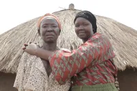 Deux femmes debout devant une hutte avec un toit de paille, regardent la caméra. La femme plus à droite sourit en serrant l’autre femme dans ses bras.