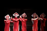 Sur une scène sombre se trouvent cinq danseuses de ballet vêtues de costumes rouges fluides aux genoux et coiffées de chapeaux carrés blancs. Elles regardent vers l'avant, les mains en prière.