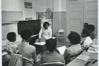Une photo en noir et blanc d’une femme noire assise sur un banc de piano, tournée aux trois quarts vers un groupe d’élèves noirs. Les élèves sont assis en demi-cercle sur des chaises pliantes en métal et tiennent des cahiers. Derrière eux se trouve une table sur laquelle sont posés deux stylos, un cahier dont la première page est remplie d’écriture et un morceau déchiré d’une affiche.