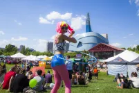 Une danseuse de hula hoop avec une perruque rose et jaune fluorescent et des leggings roses marche dans une foule de personnes assises sur un champ d'herbe devant un grand bâtiment en verre.
