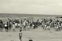 Une photographie en noir et blanc d’une foule de personnes, la plupart debout, sur une plage.