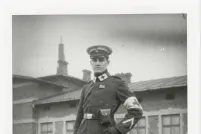 Une photo en noir et blanc d’un homme vêtu d’un uniforme d’infirmier militaire, arborant un brassard blanc orné d’une croix. De chaque côté du col se trouve un écusson blanc sur lequel figure une grande croix. L’homme regarde droit vers l’appareil photo, le visage impassible. Un bâtiment se trouve derrière lui.