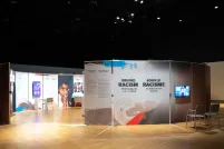 Des panneaux verticaux sont disposés en cercle dans une pièce faiblement éclairée. Une ouverture permet aux gens d'entrer dans le cercle, et l'intérieur du cercle est bien éclairé. À côté de l'ouverture, on peut lire sur un panneau les mots « Sous le racisme : Un défi pour nos pensées ».