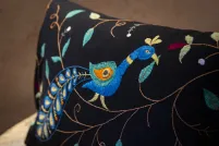 Photo en gros plan d’un coussin décoratif rectangulaire noir orné d’une broderie détaillée représentant un paon stylisé au cou bleu incurvé, à la crête bleu sarcelle et à la plume aux motifs oculaires orange et bleu sarcelle, posé sur un banc en bois. Le motif comprend également des surpiqûres marron en forme de vigne, des feuilles bleu sarcelle et sauge, ainsi que de petits motifs floraux bordeaux et blancs disséminés sur le tissu noir.