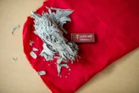 Un petit paquet de sauge séchée repose sur un tissu rouge vif, entouré de fragments de feuilles éparpillés. À côté se trouve une petite boîte d’allumettes rectangulaire portant l’inscription « Mother Earth Tobacco ». Ces objets sont posés sur une surface en bois clair.