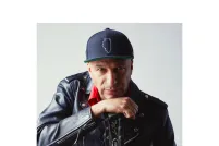 Tom Morello regarde intensément la caméra tout en s’appuyant sur sa main qui tient une guitare par le manche. Il porte une veste en cuir noire, une chemise à col rouge et une casquette.