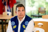 A man wearing a blue vest with flower beadwork, seated with his hands resting on a table.
