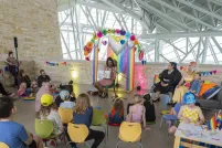 Une artiste drag fait la lecture à un groupe d'enfants et d'adultes assis sur des chaises colorées, dans un lieu lumineux de style industriel décoré de banderoles arc-en-ciel et de guirlandes, à l'occasion d'une heure du conte sur le thème de la Fierté.