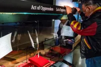 Un homme aux longs cheveux tressés et à la tête penchée se tient devant une vitrine où se trouvent deux plumes.