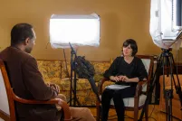 Un homme et une femme sont assis face à face dans des chaises. On voit un microphone sur pied entre eux, ainsi que de grandes lampes de chaque côté de la chaise de la femme. Les lampes sont pointées vers l’homme.