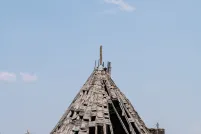 Une grande structure en bois patinée par les intempéries, en forme de tipi, se dresse dans un champ d’herbe sèche sous un ciel bleu limpide. L’ossature en bois de la structure est en grande partie apparente; de nombreux bardeaux manquant ou tombant, laissant apparaître le squelette nervuré en dessous.