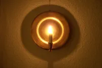 La lueur d’une chandelle sur une applique murale ronde en laiton; son réflecteur brille d’un éclat doré et projette une ombre circulaire sur un mur texturé.
