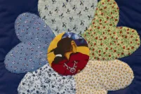 Gros plan sur une fleur sur une courtepointe. Chacun des pétales est un tissu fleuri différent, tandis qu’au centre de la fleur se trouve un tissu sur lequel est imprimé le profil d’une femme autochtone.