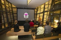 Un petit groupe de personnes assises sur des bancs en bois dans un théâtre fait face à un écran montrant une femme âgée assise dans un fauteuil. Un homme vêtu d’une chemise jaune se tient à droite de l’écran et tient un microphone.