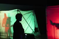 La silhouette d’une personne debout dans le noir devant des structures en tissu rouge et vert illuminées de l’intérieur et contenant des silhouettes ombragées.