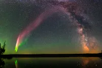 Un arc éclatant d’aurores rose-rougeâtre et vertes se dessine dans le ciel nocturne étoilé, le long du cœur de la Voie lactée, au-dessus d’un lac sombre bordé d’arbres dont la surface calme reflète ces lumières.