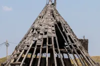 Une grande structure en bois patinée par les intempéries, en forme de tipi, se dresse dans un champ d’herbe sèche sous un ciel bleu limpide. L’ossature en bois de la structure est en grande partie apparente; de nombreux bardeaux manquant ou tombant, laissant apparaître le squelette nervuré en dessous.
