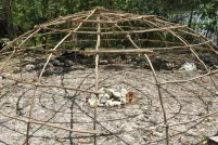 Une charpente en forme de dôme, constituée de perches de bois entrelacées, avec au centre un tas de pierres. La structure est entourée d’arbres avec de l’eau en arrière-plan.
