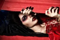 Une vampire vêtue d’une robe rouge en velours regarde à travers une boîte en bois rouge qu’elle entrouvre avec ses longs ongles noirs.