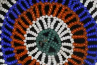 Une photo en gros plan d’un motif circulaire en perles sur fond noir. Le motif se compose d’anneaux concentriques de perles turquoise, blanches, orange et bleu vif, disposées en forme de fleur ou de soleil.