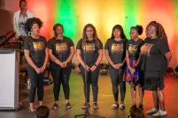 Un groupe de six artistes noires debout sur scène avec un rétroéclairage coloré.
