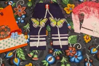 Des pièces de vêtements artisanaux de style autochtone sont posées sur un morceau de papier artisanal orné de fleurs et de baies. Les vêtements comprennent une chemise orange, un pantalon et une tunique rose décorée de tissus, d'appliques et de rubans colorés. La tunique comprend un châle à plumes sur les épaules.