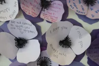 Des fleurs à trois pétales en papier de différentes nuances de violet sont regroupées dans une œuvre murale. Des noms et des messages y sont inscrits.