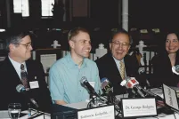 Un homme souriant vêtu d’une chemise bleu clair est assis à une longue table devant un groupe de microphones. Assis de part et d’autre de lui, d’autres personnes en complet sont également tournées vers les microphones et sourient. Les plaquettes nominatives le long de la table indiquent, de gauche à droite, Murray Billet, Robinson Koilpillai et Dr George Rodgers, avec des affiliations illisibles sous les noms. Une autre plaquette, renversée sur le côté, indique Delwin Vriend sans affiliation.